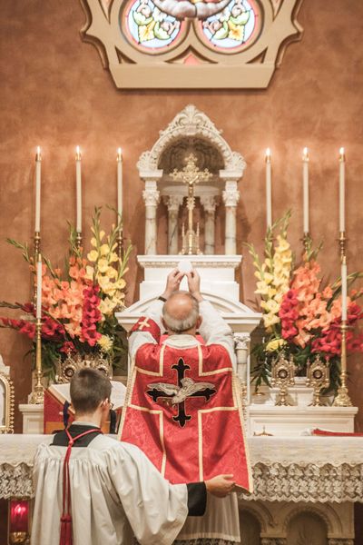 The priest, in red vestments, raises the Sacred Host at the high altar during Mass, with an altar server beside him and orange and red flowers on the altar.
