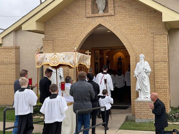Altar servers and faithful in front of the arched chapel entrance under a processional canopy, with a white statue of Christ beside the door.