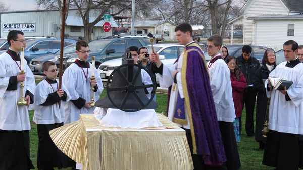 A priest in a purple stole blesses a large bronze bell resting on a draped table outdoors, surrounded by altar servers holding candles.