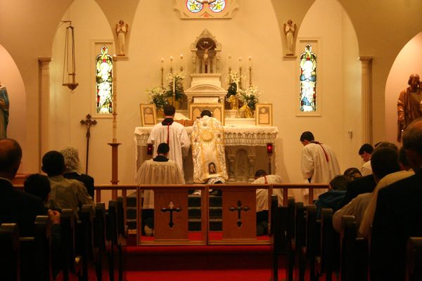 View from the pews toward the altar, where a priest in a white and gold chasuble offers Mass with servers kneeling, Easter lilies on the altar.