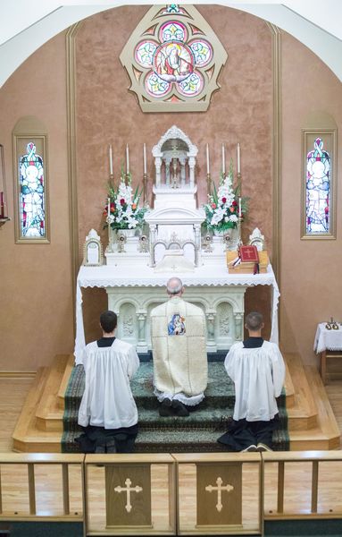 A priest in a white Marian chasuble kneels at the foot of the white high altar between two kneeling altar servers.