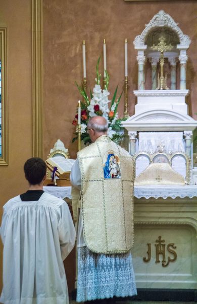 A priest in a white chasuble embroidered with an image of the Madonna and Child stands at the altar beside the open missal while an altar server waits nearby.