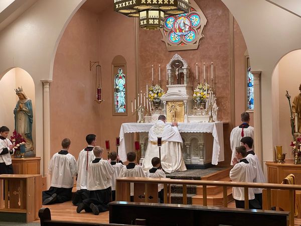 Wide view of the sanctuary with a priest at the high altar and altar servers kneeling on the steps, statues and stained glass on either side.