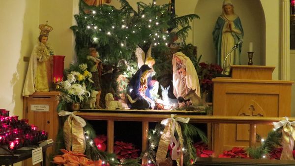 A lit Nativity scene beneath a decorated Christmas tree with red poinsettias, flanked by statues of the Blessed Virgin and a saint.