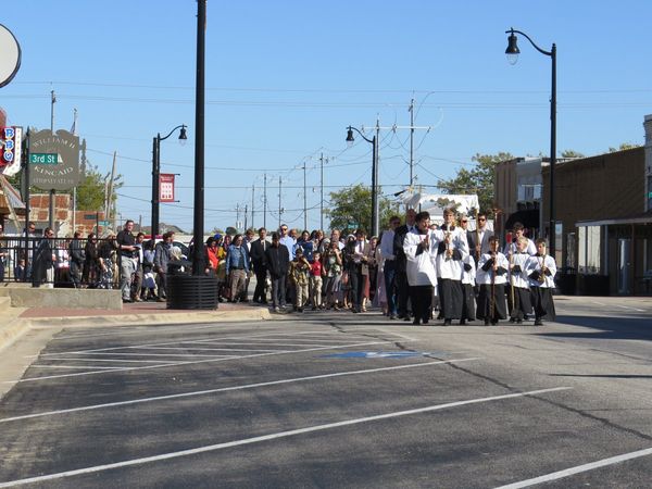 Cross bearer, candle bearers and a canopy lead a long procession of faithful down a town street on a sunny day.