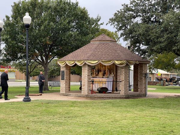 An octagonal gazebo on a green lawn, decorated with white drapery and an altar with candles, shaded by large trees.
