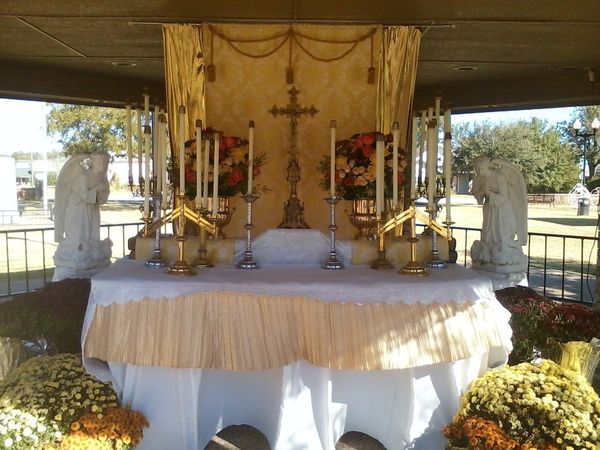 An outdoor altar draped in white and gold with tall candles, a crucifix and gold flowers, with potted yellow mums in front.