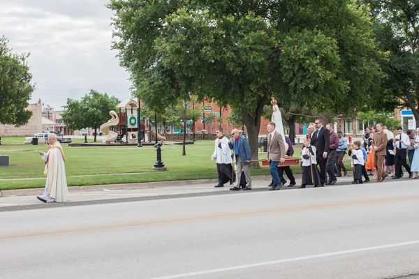 A priest in a cope leads men carrying the crowned statue of Our Lady of Fatima on a wooden litter along a park walkway, followed by the faithful.