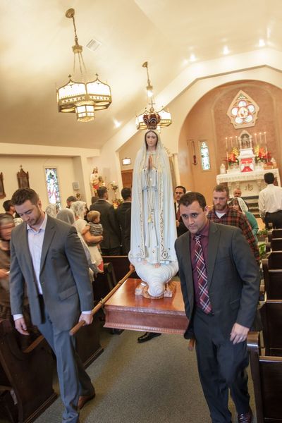 Two men carry the Our Lady of Fatima pilgrim statue on a wooden litter down the chapel aisle, with the faithful in the pews and the altar behind.
