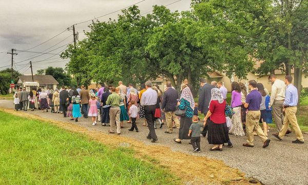 A long line of faithful, many women in veils, walking along a gravel road behind the canopy in a Corpus Christi procession.