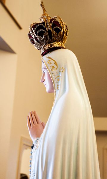 Close side view of the crowned Our Lady of Fatima statue, hands folded in prayer, in a white mantle trimmed with gold.