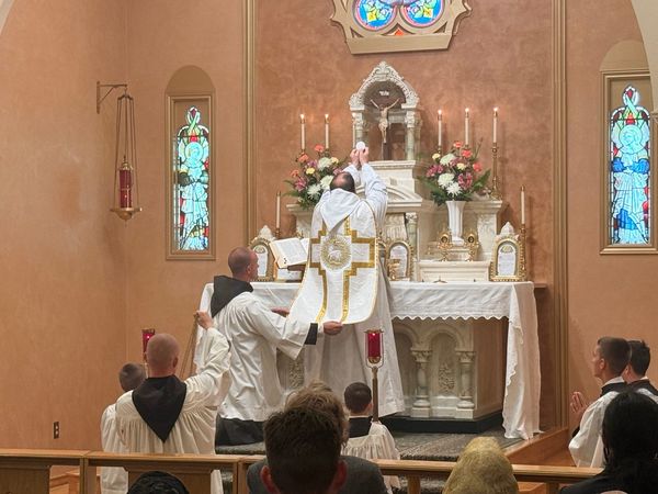 A priest in a white chasuble with a gold cross raises the Host at the white high altar as two men in white hold the edge of his chasuble, with flowers, candles and stained glass windows on either side and the faithful in the pews.