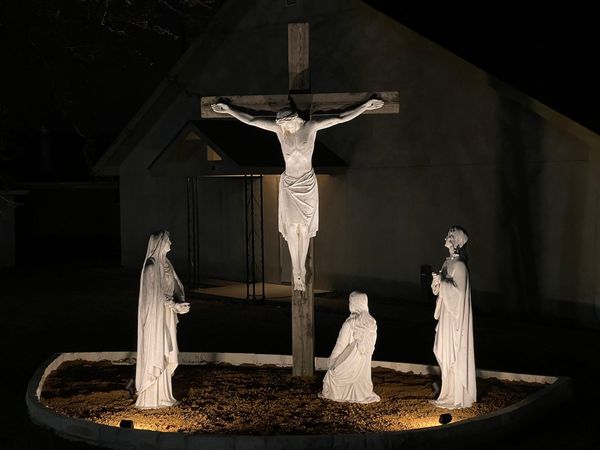 A life-size outdoor crucifix lit at night, with statues of the Blessed Virgin, St. Mary Magdalene kneeling and St. John at the foot of the cross in front of the chapel.