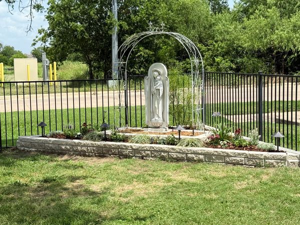 A white statue of Our Lady holding the Child Jesus under an ornate white metal arch in a raised stone flower bed with path lights, an iron fence behind.